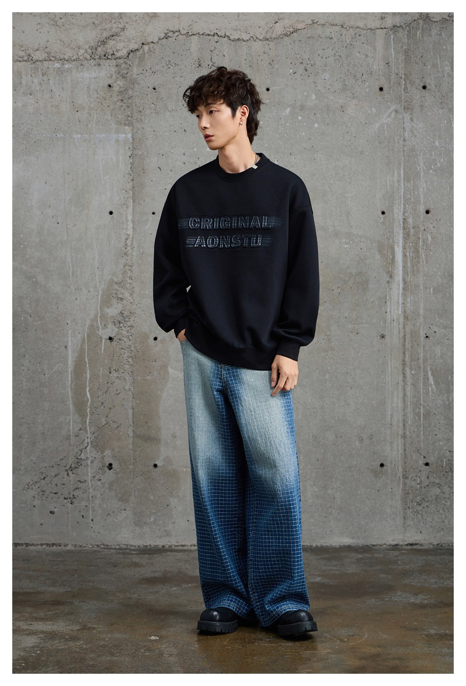 Model wearing a black crewneck sweatshirt with raised lettering and loose blue plaid trousers.