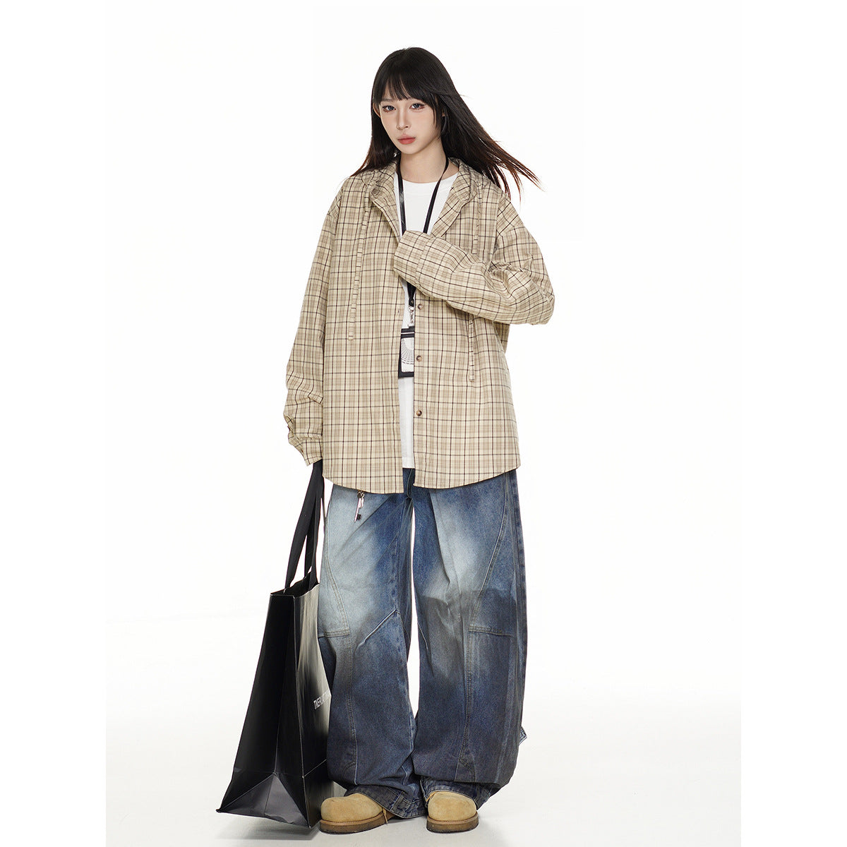 Model wearing a beige plaid overshirt and wide blue jeans, carrying a black tote bag.