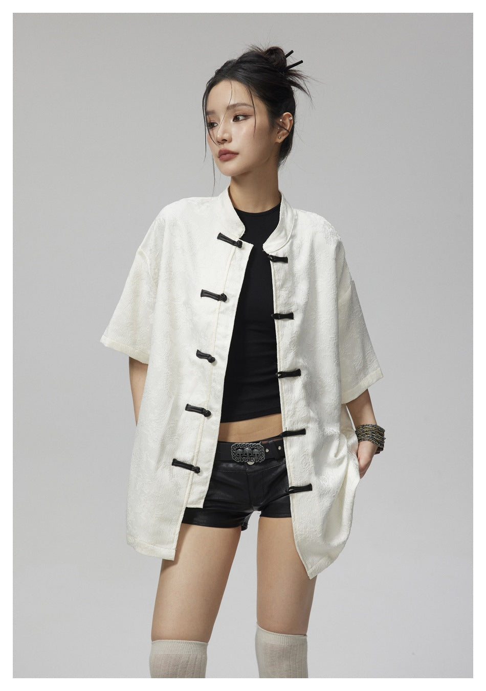 Model wearing an ivory short-sleeve mandarin-collar jacket with black frog closures over a black top and shorts.