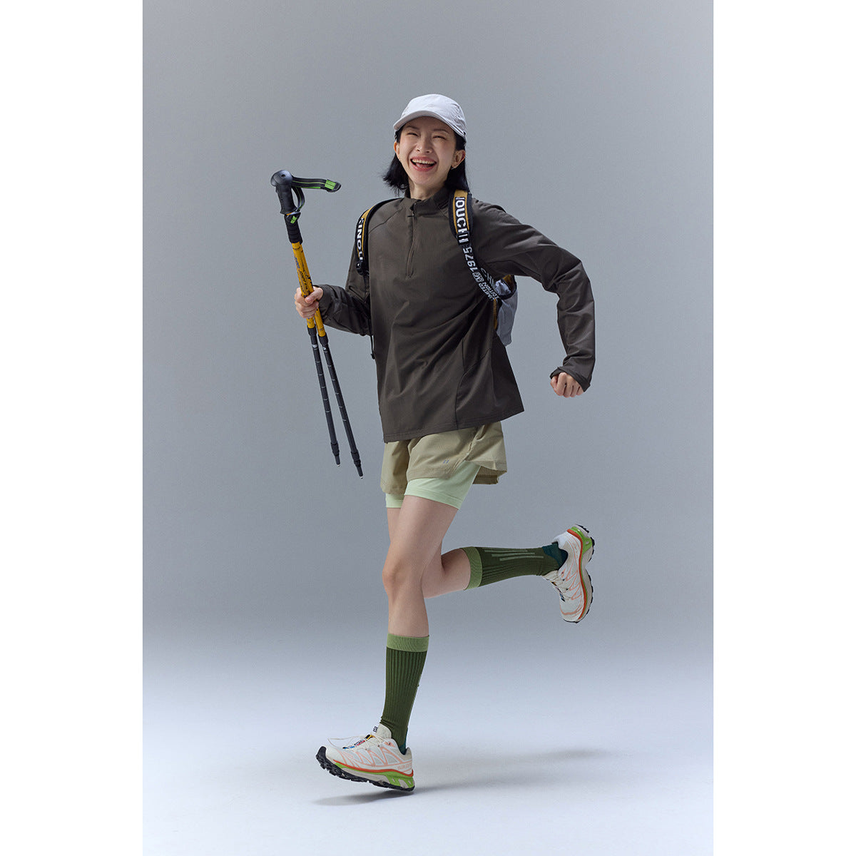 A woman models a dark brown zip-neck top with layered khaki shorts, a white cap and green socks.