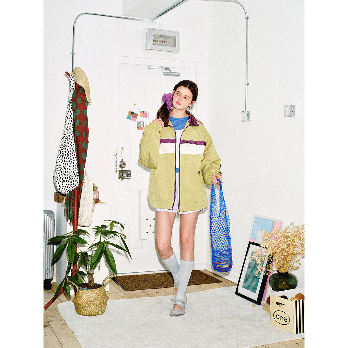 Model poses in a khaki track jacket with purple-and-white chest stripes and striped sleeves, holding a blue mesh bag.