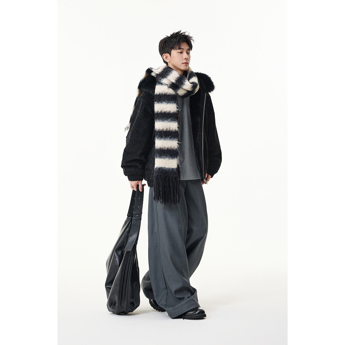 Man wearing a black hooded jacket with plush trim, a black-and-cream striped scarf, and loose gray trousers.