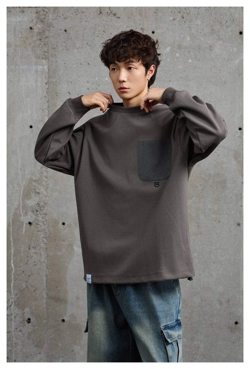 Model adjusts the collar of a charcoal gray oversized sweatshirt with a dark chest pocket.