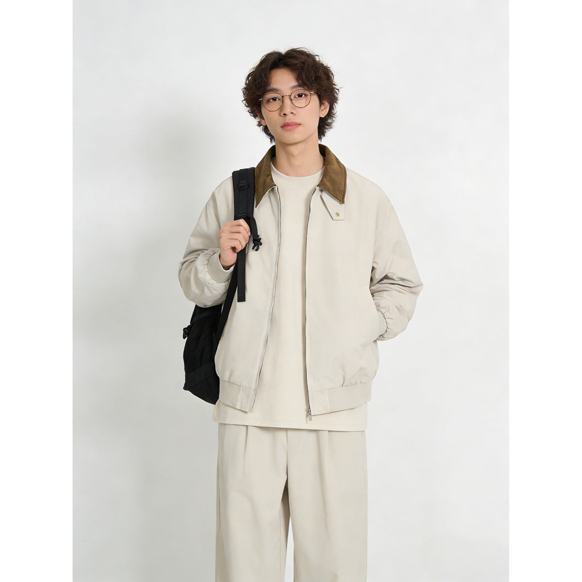 Model wearing a light cream zip jacket with a brown collar, paired with cream trousers and a black backpack.