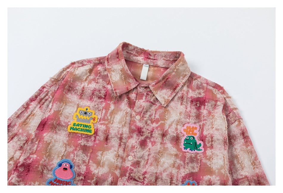 Upper front of a pink, cream and tan distressed plaid button-up shirt with colorful embroidered character patches.
