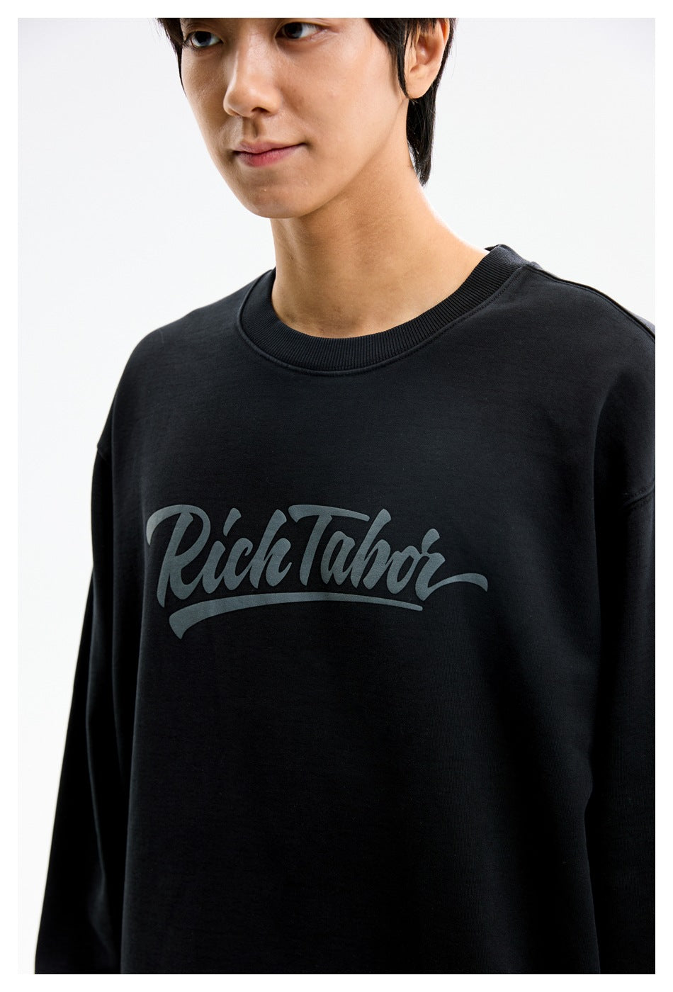 Close-up of a model wearing a black crewneck sweatshirt with gray script lettering across the chest.