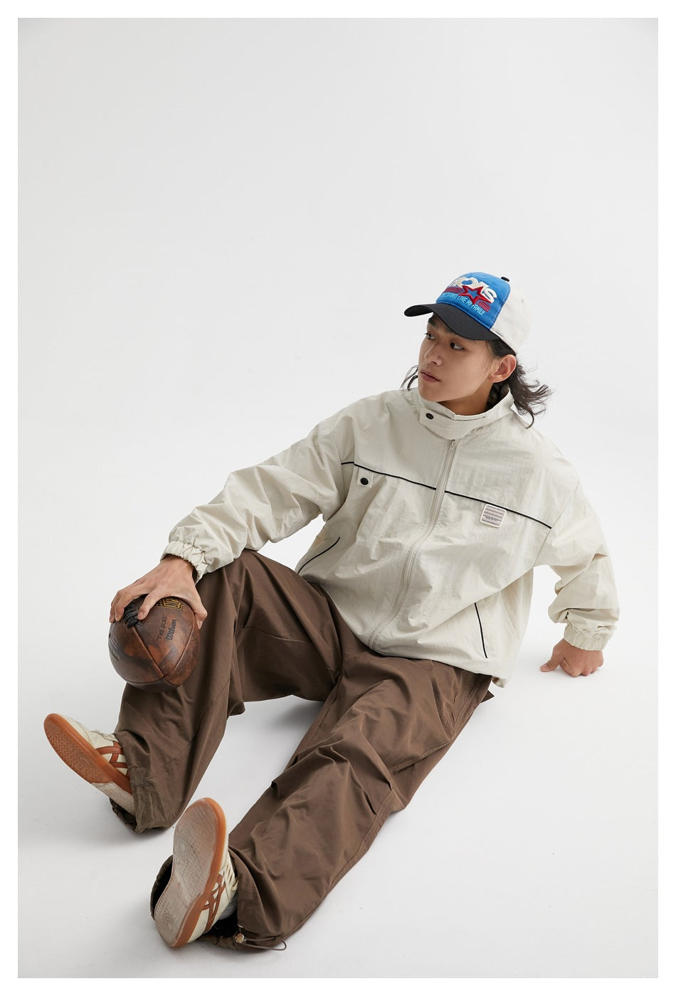 Seated model wearing an ivory zip jacket with black piping and loose brown trousers, holding a football.
