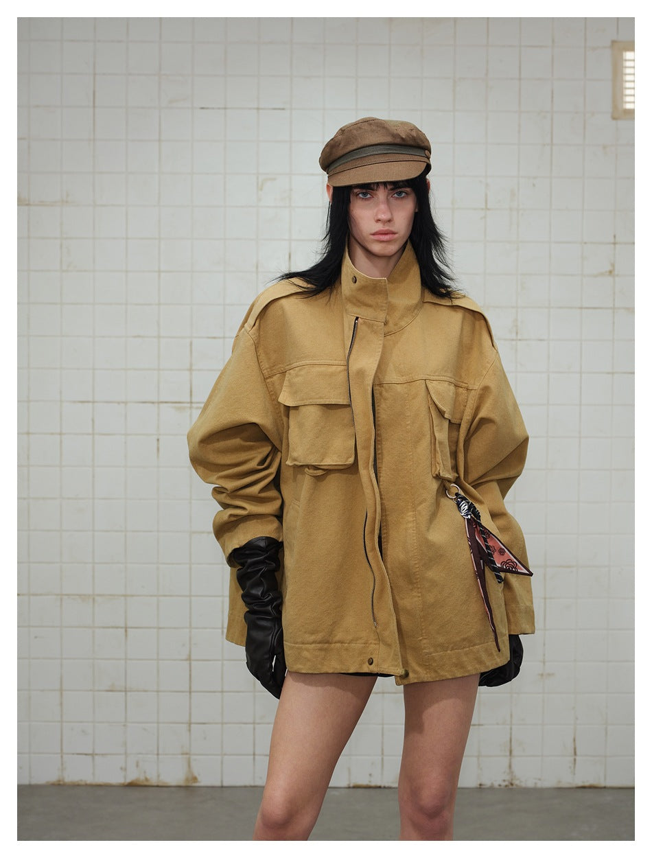 Model wearing oversized brown stand-collar jacket with flap pockets and scarf charm, with cap and black gloves