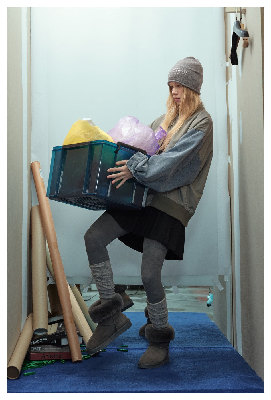 Model carrying a bin while wearing a gray-green bomber jacket with blue denim sleeves and a black skirt.