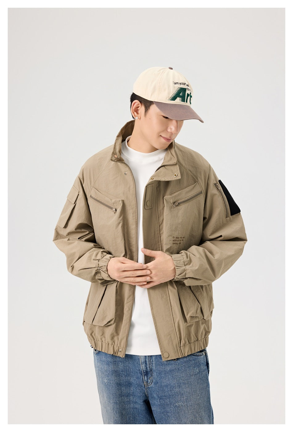 Model looking down while wearing a khaki utility jacket with multiple pockets, a white shirt, and a cream cap.