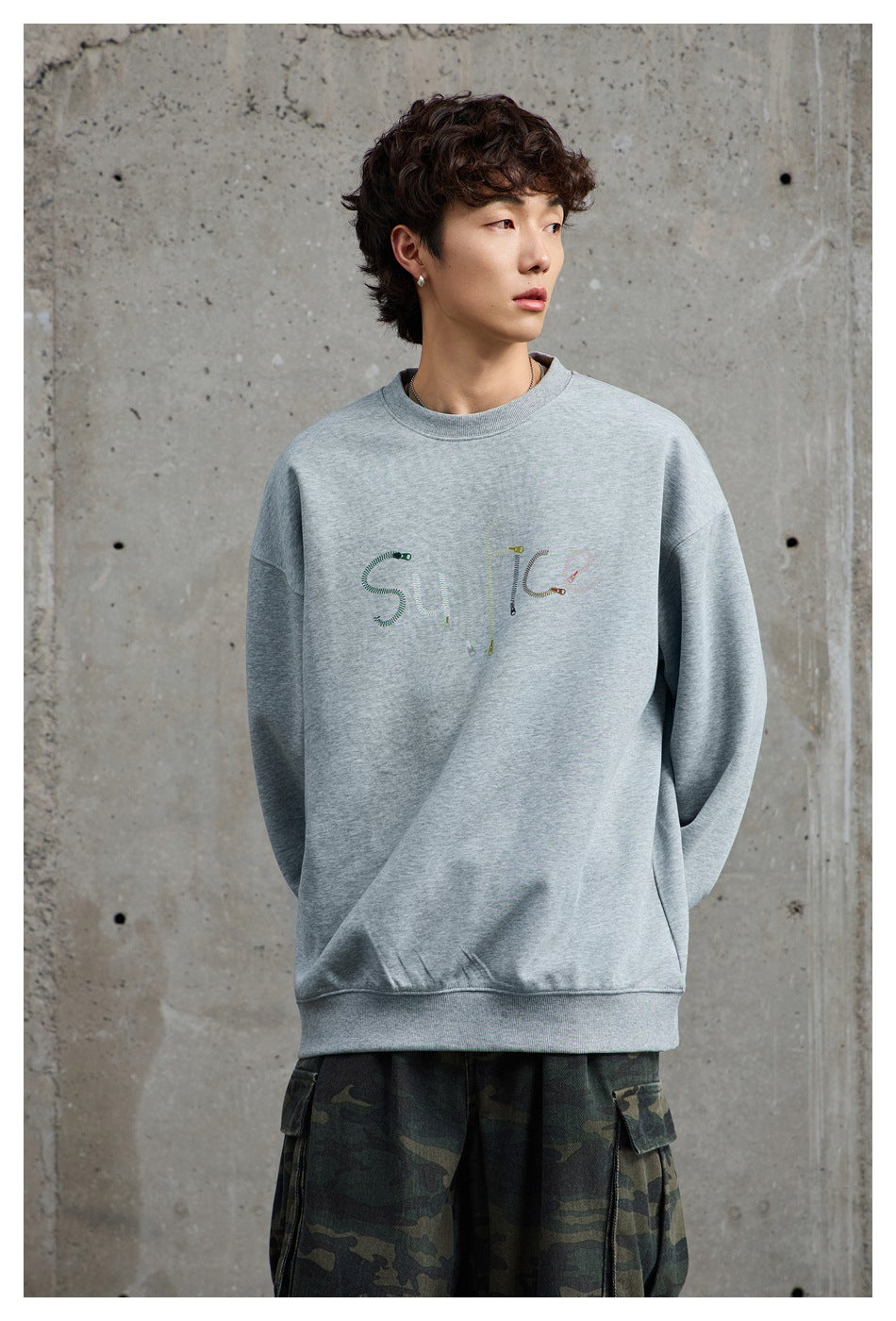 Model in a light gray crewneck sweatshirt with subtle multicolored chest lettering and dark camouflage cargo pants.