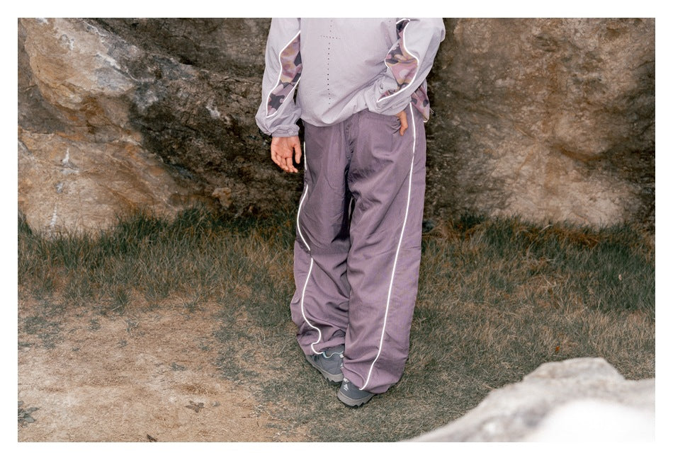 Rear view of a model wearing loose muted lavender pants with white side piping outdoors.