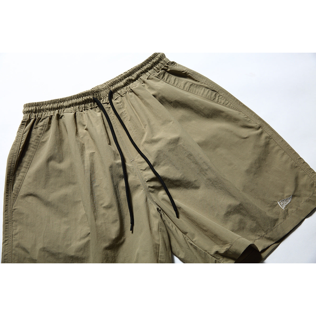 Front close-up of olive-khaki shorts with an elastic drawstring waist, side pockets and a small embroidered leg emblem.