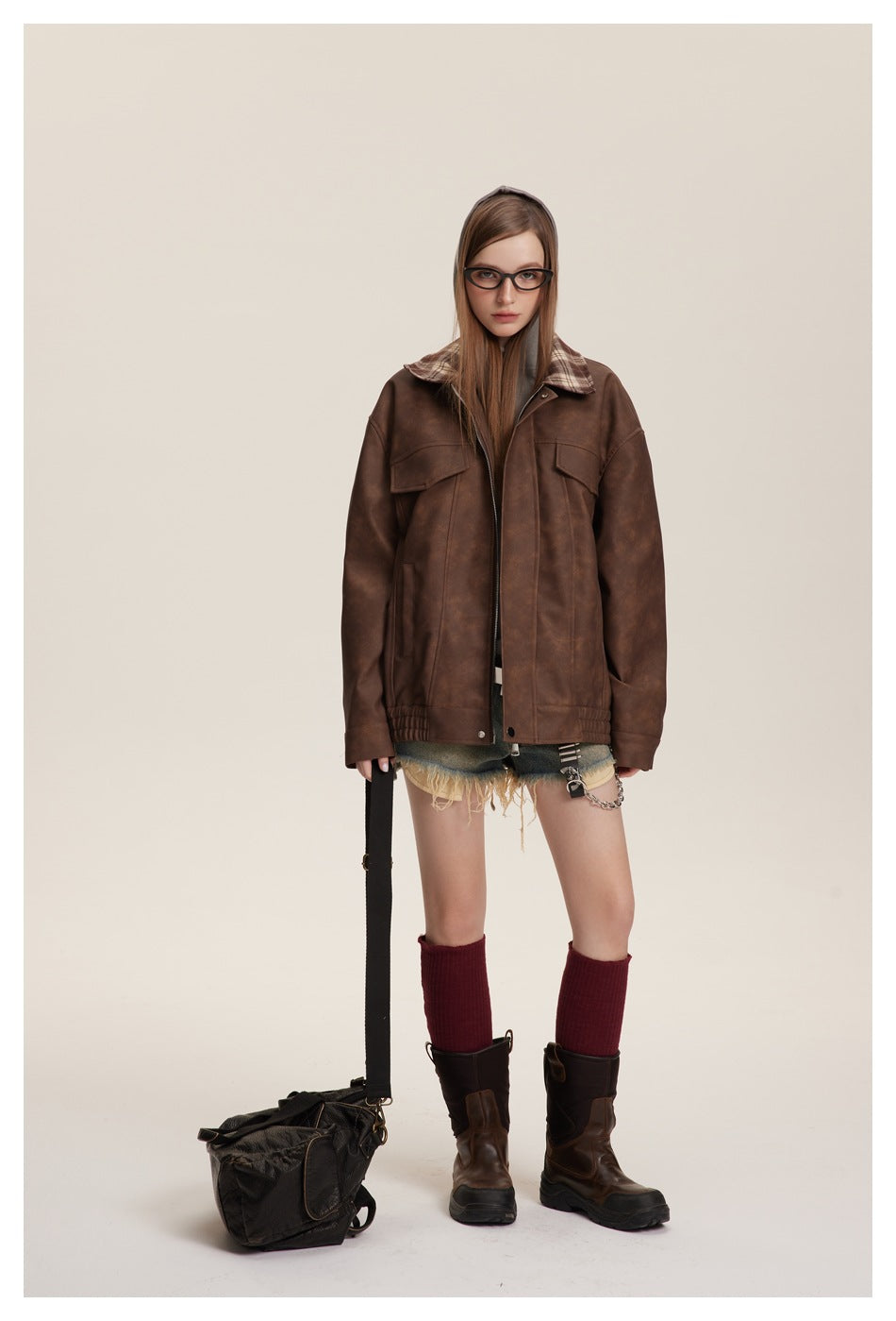 Model wearing an oversized brown jacket with plaid collar, gray hood, flap pockets, distressed shorts and boots.