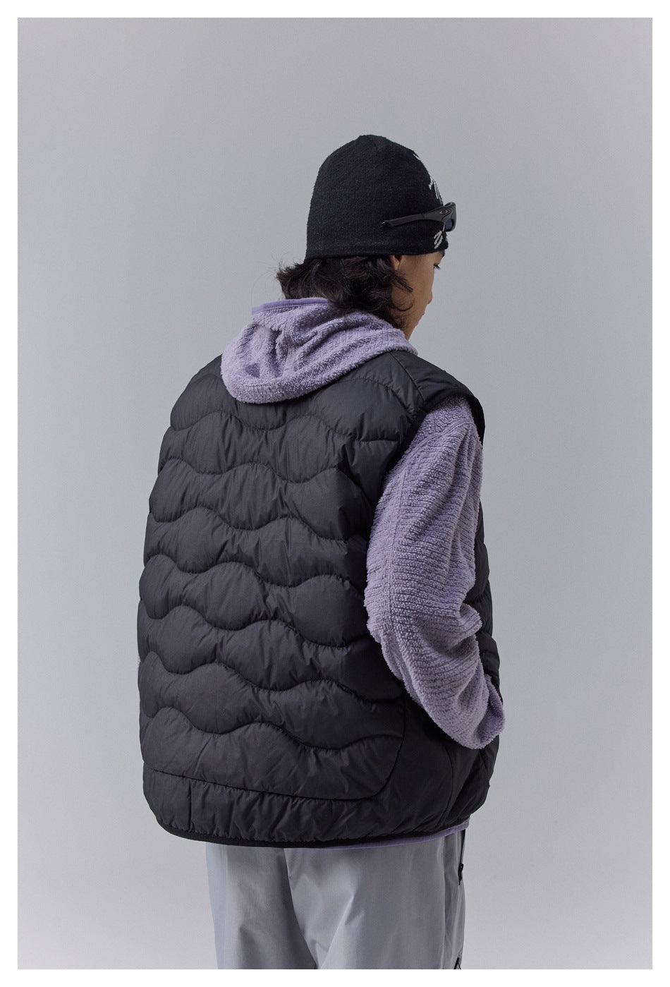 Rear view of a model wearing a black quilted vest over a lavender hooded fleece top.