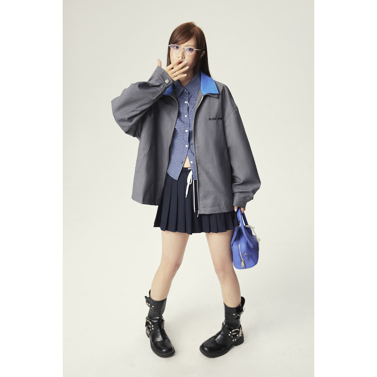 Full-length model wearing a gray zip jacket with a vivid blue collar, navy pleated skirt, and black boots.