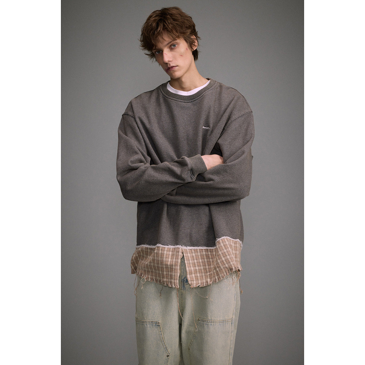 Man modeling a charcoal oversized sweatshirt with a distressed plaid hem and loose pale jeans, arms crossed.