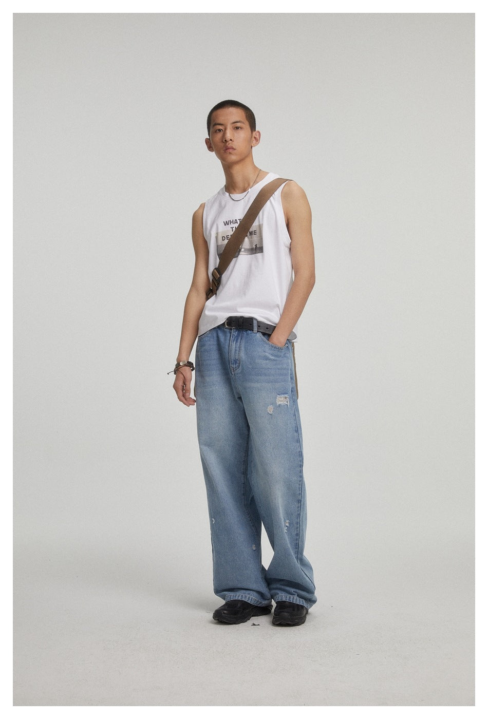 Model wearing a white graphic sleeveless shirt and loose distressed blue jeans, with a brown crossbody strap.