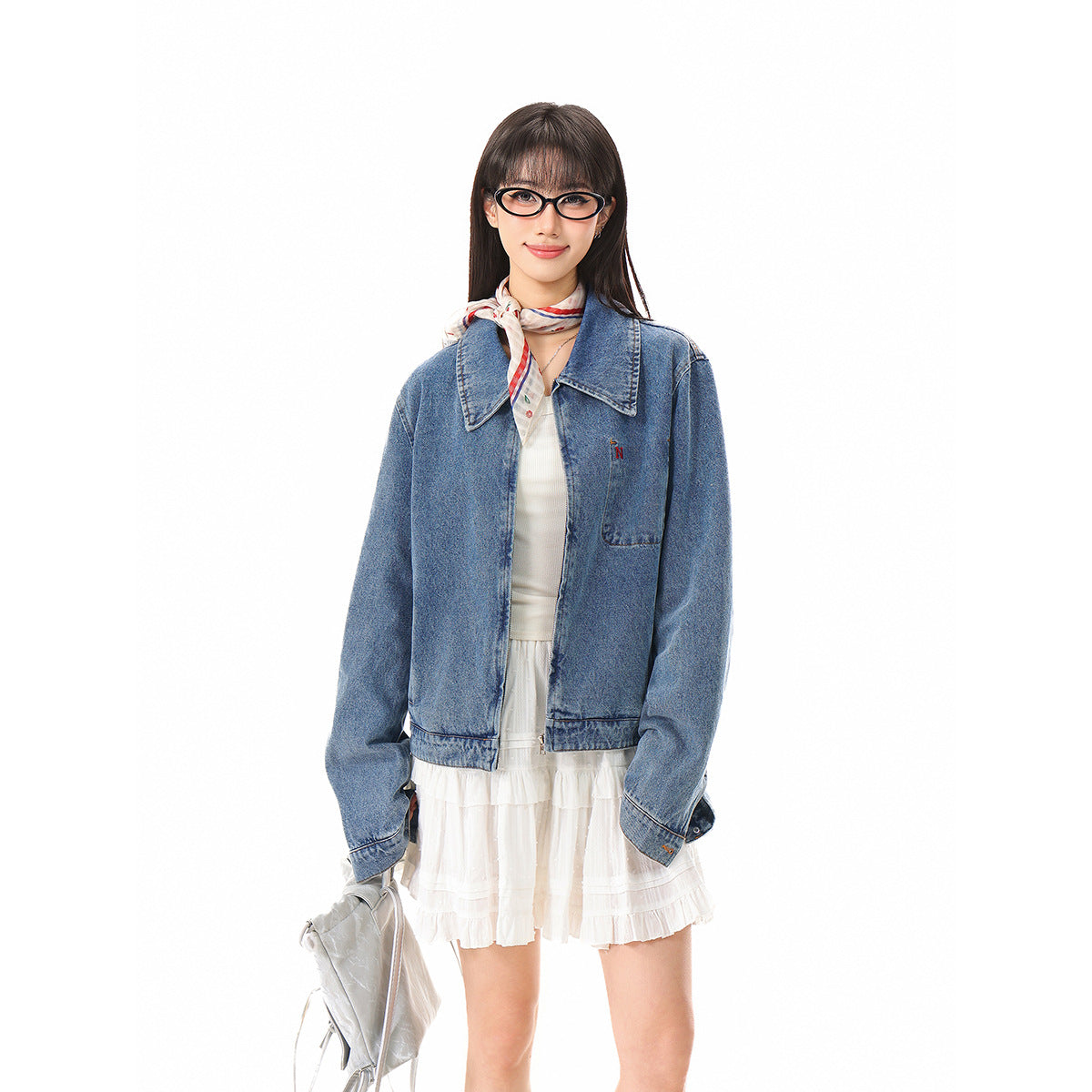 Front view of a model in an oversized blue denim jacket over a white ruffled dress, with a silver bag at her side.