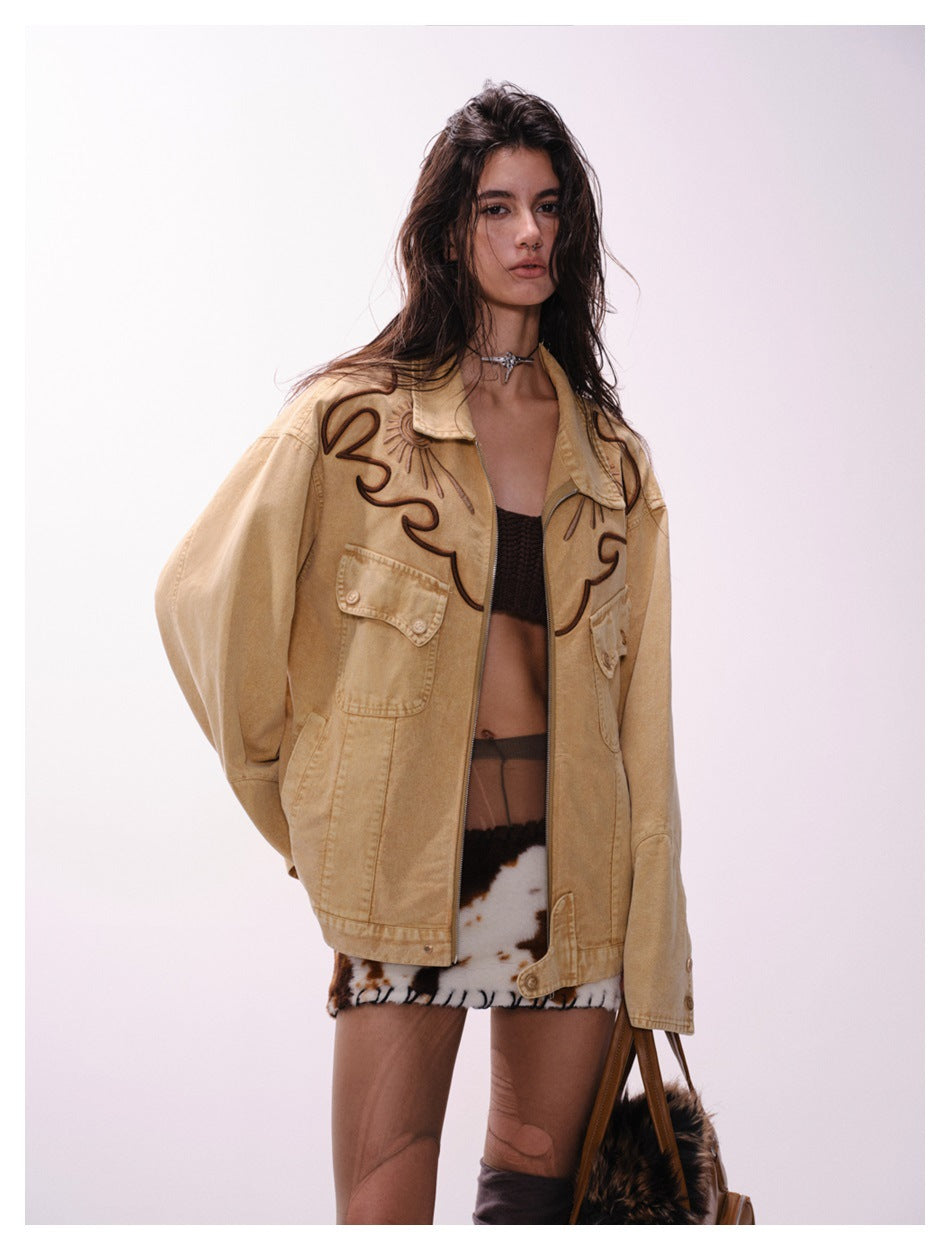 Model wearing an oversized tan jacket with dark embroidered motifs and flap pockets, styled with a black crop top and