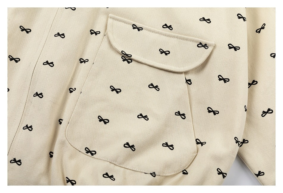 Close-up of a beige garment with repeating black bow motifs, a large patch pocket, and a wide folded collar.