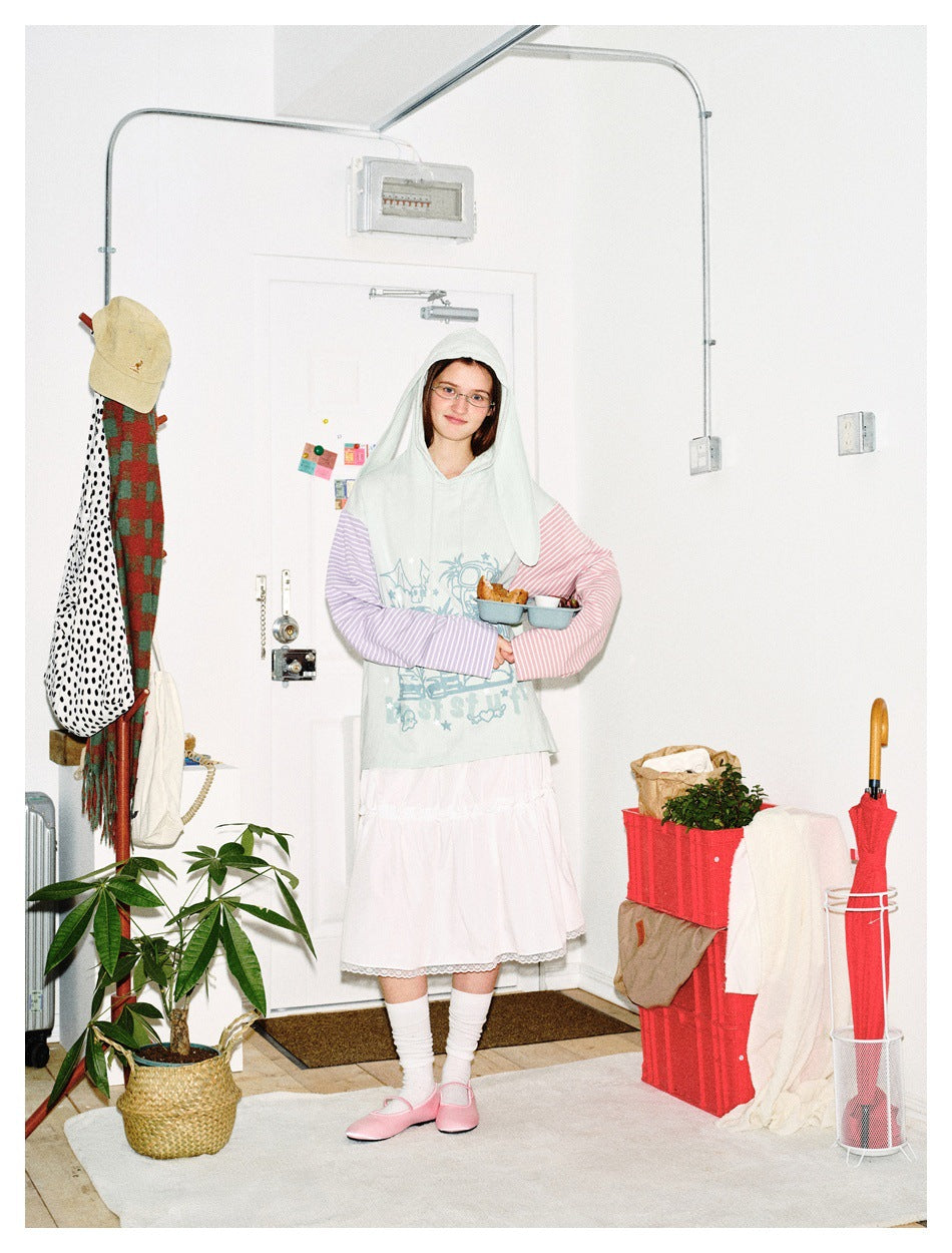 Full-length model wearing a pale mint graphic hoodie with long hood panels and pink and lilac striped sleeves.