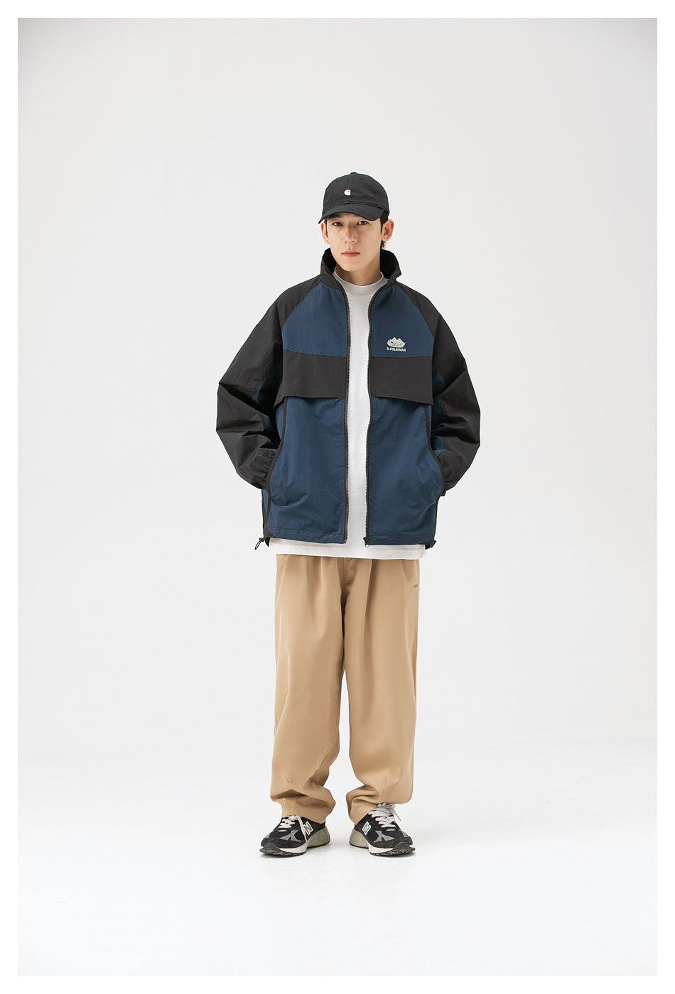 Model wearing navy and black zip jacket over a white tee with khaki trousers and a black cap, front view