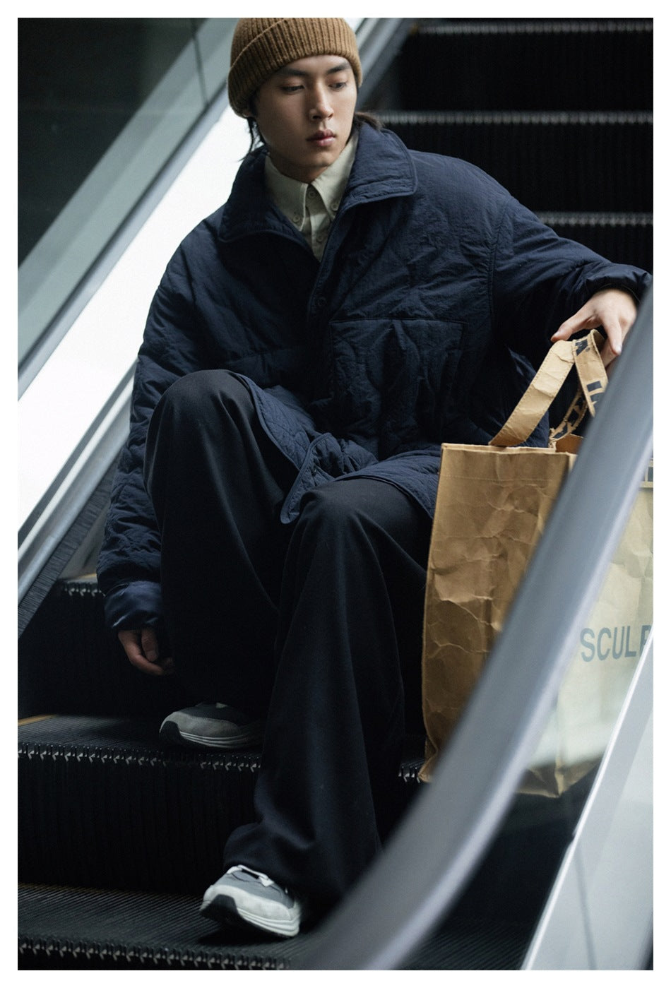 Seated model wearing a dark navy quilted jacket and brown ribbed beanie on an escalator.