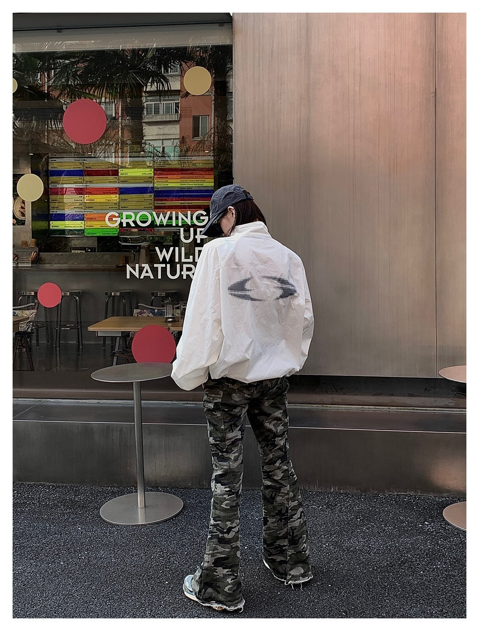 Rear view of a model wearing a white jacket with a large dark back emblem and camouflage flared pants.