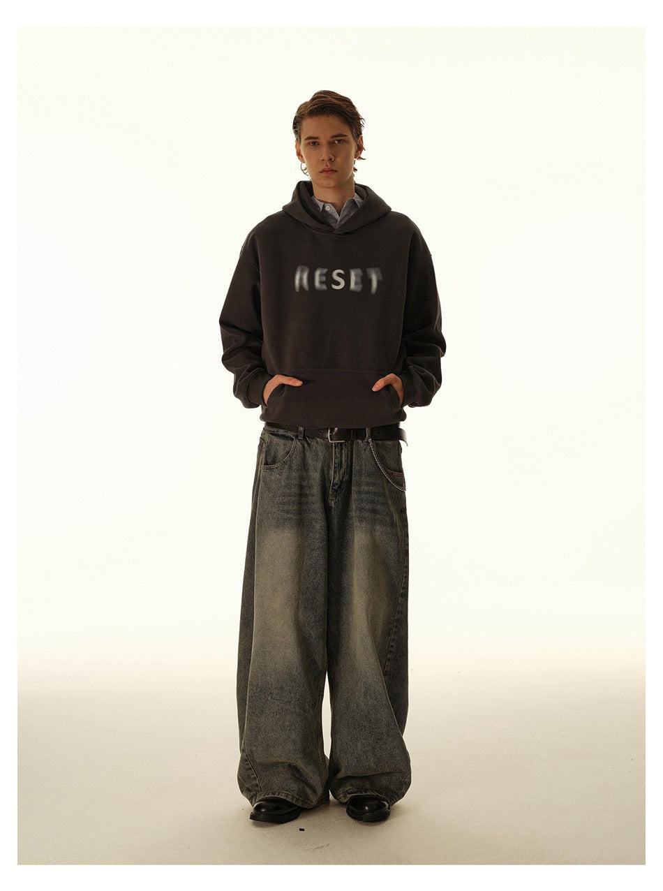Front view of a model wearing a dark brown hoodie with blurred chest lettering and wide gray-black jeans.