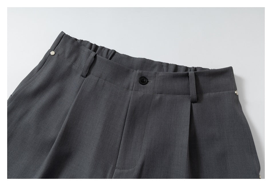 Close view of dark charcoal trousers showing the front waistband, button, belt loops, pleats and pocket openings.