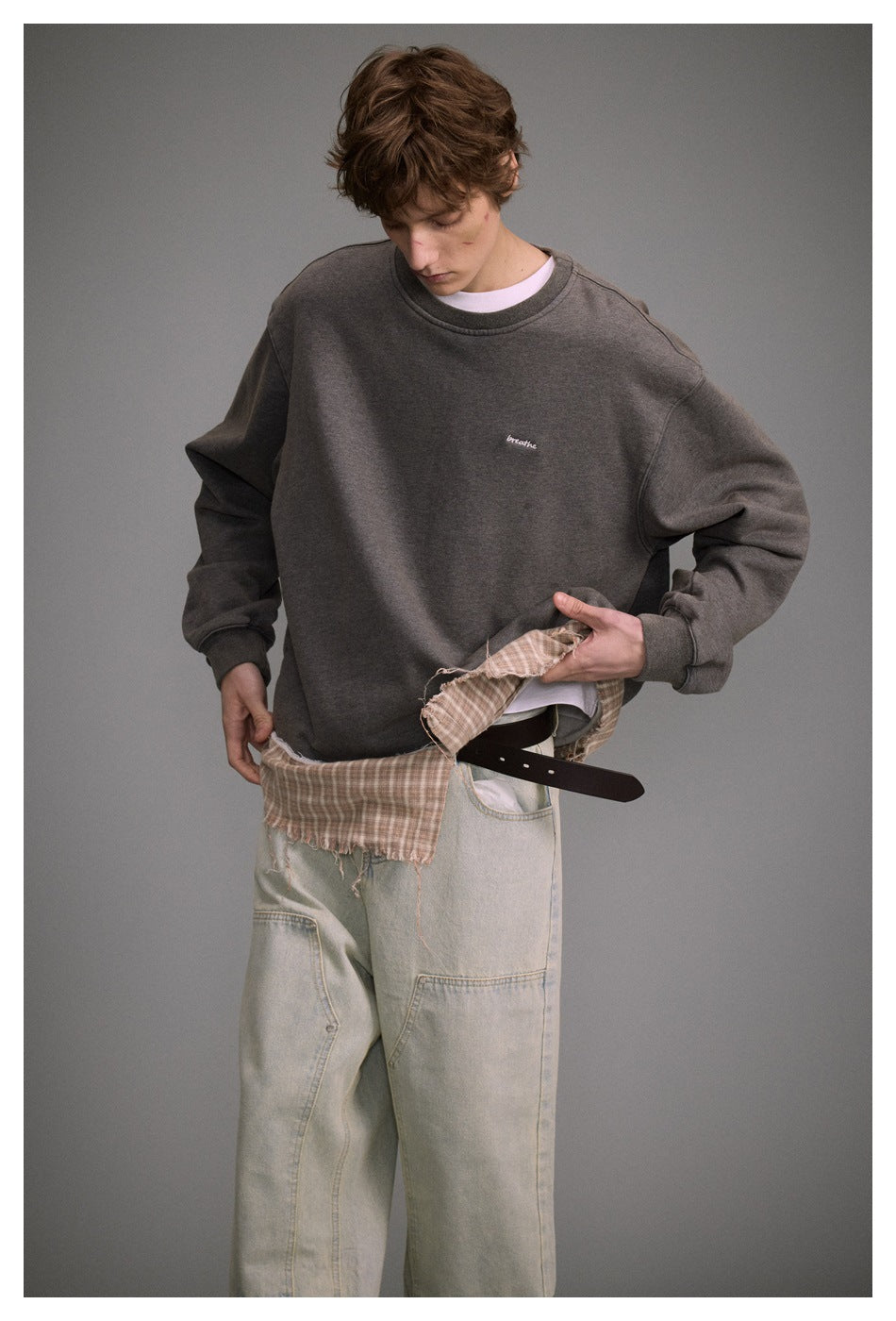 Model lifting the plaid hem panel of a gray sweatshirt over a white tee, with light wash jeans and brown belt