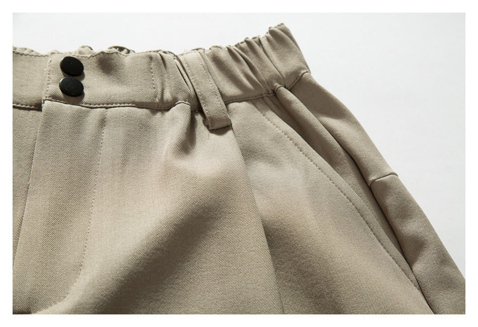 Close-up of taupe trousers showing a two-button waistband, belt loop, pleats and angled pocket opening.