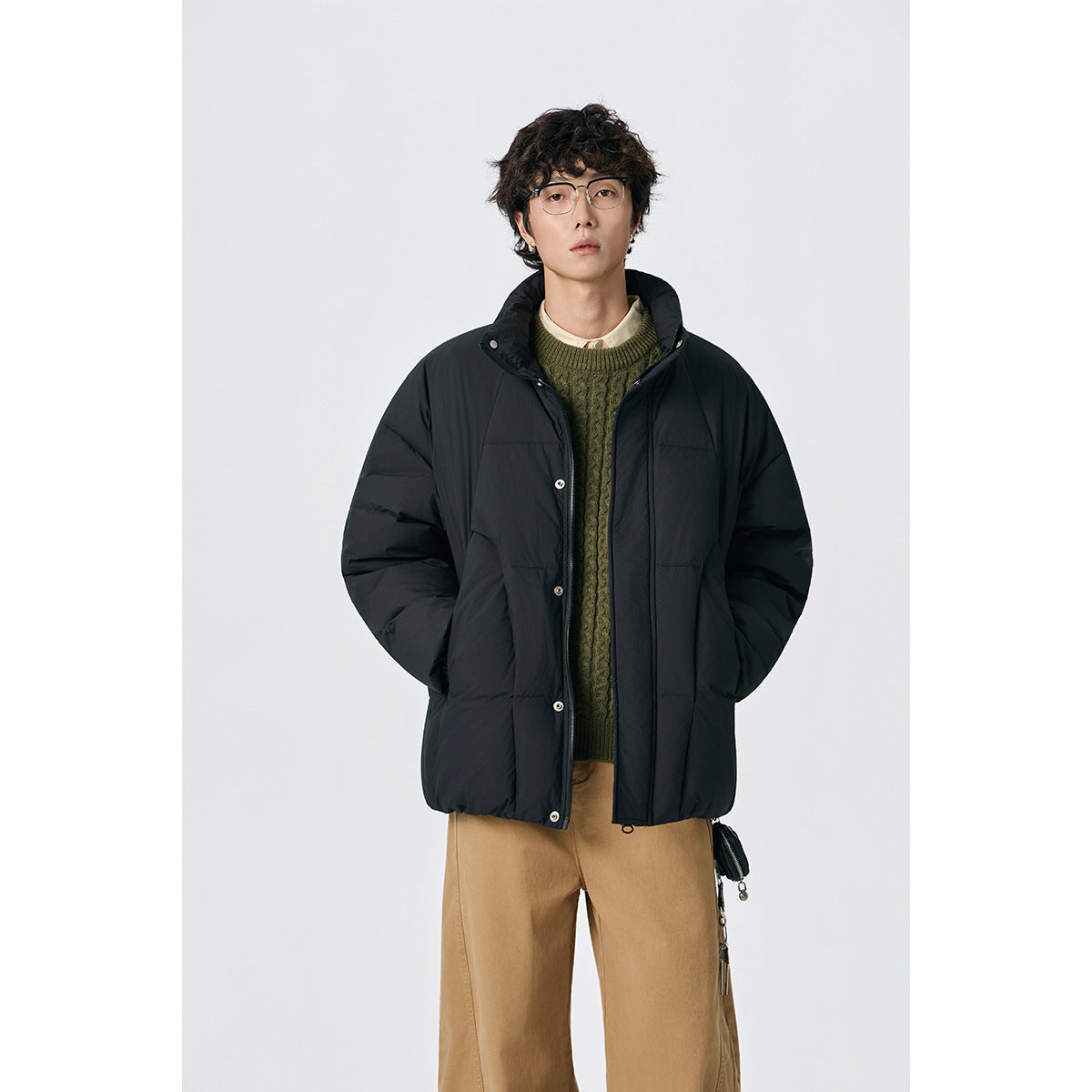 Model wearing a black padded jacket with a high collar over a green sweater and tan trousers, front view.