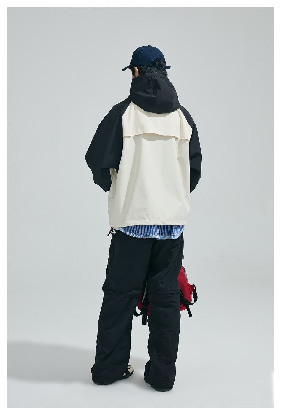 Rear view of a model wearing a cream-and-black hooded jacket with black trousers and a navy cap.