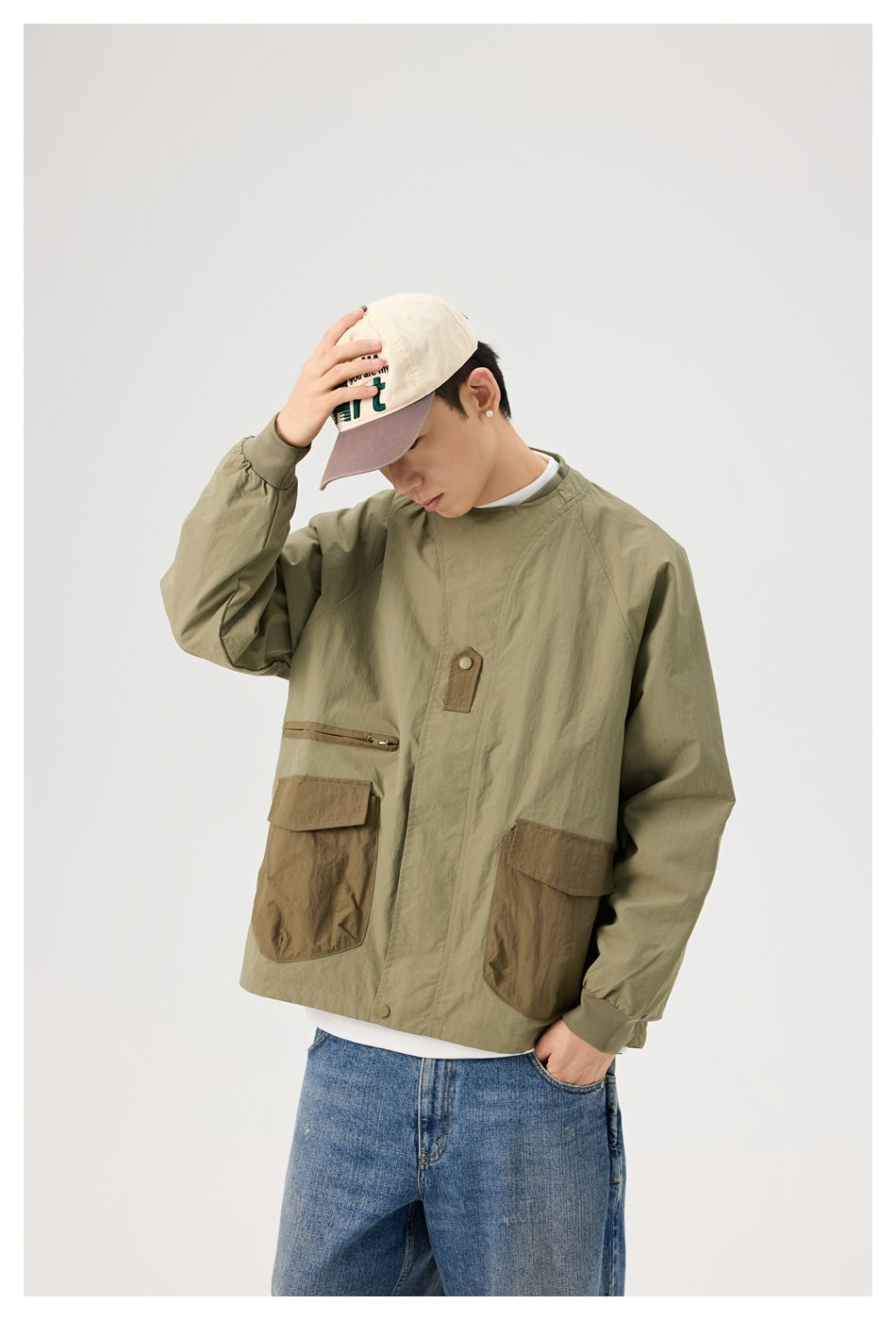 Model wearing an olive utility jacket with brown patch pockets and ribbed cuffs, shown at an angle with one hand on his cap.