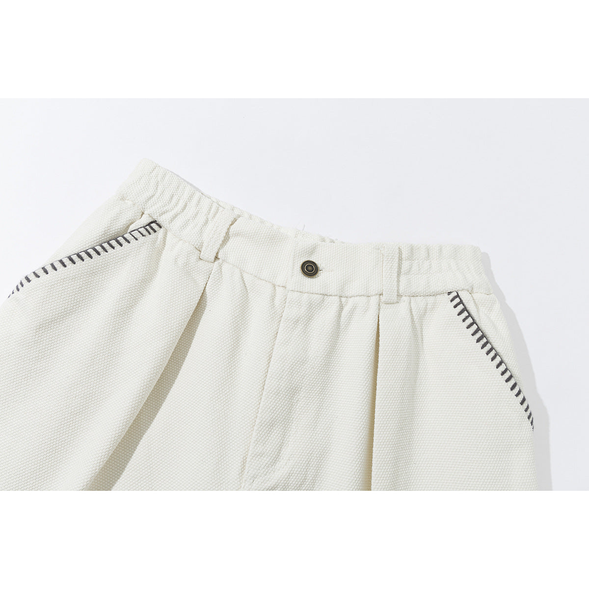 Close-up of ivory shorts showing the waistband, button, belt loops, pleats and dark contrast stitching around the pockets.