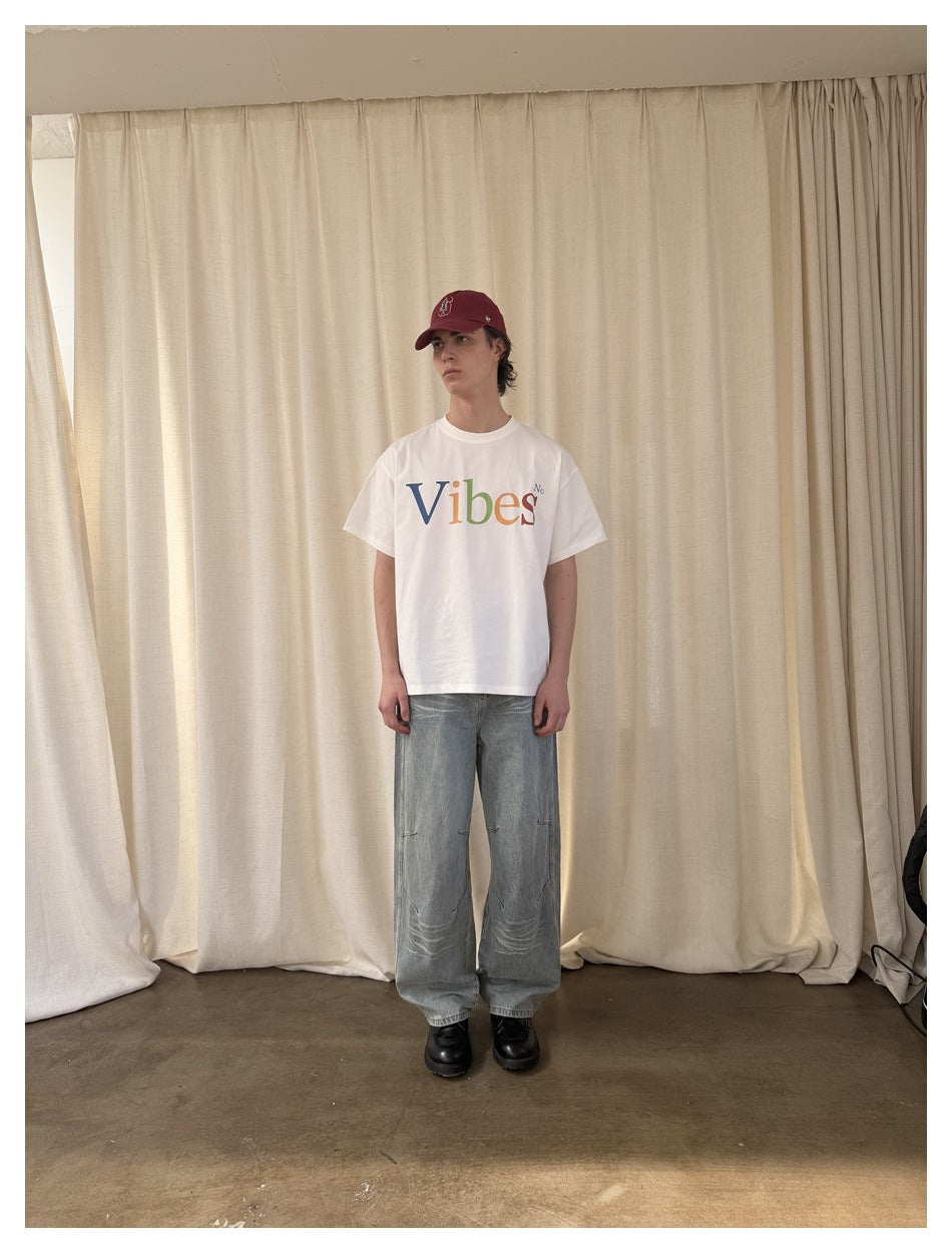 Model standing in front of a cream curtain wearing light blue wide-leg jeans, a white graphic T-shirt, red cap and black
