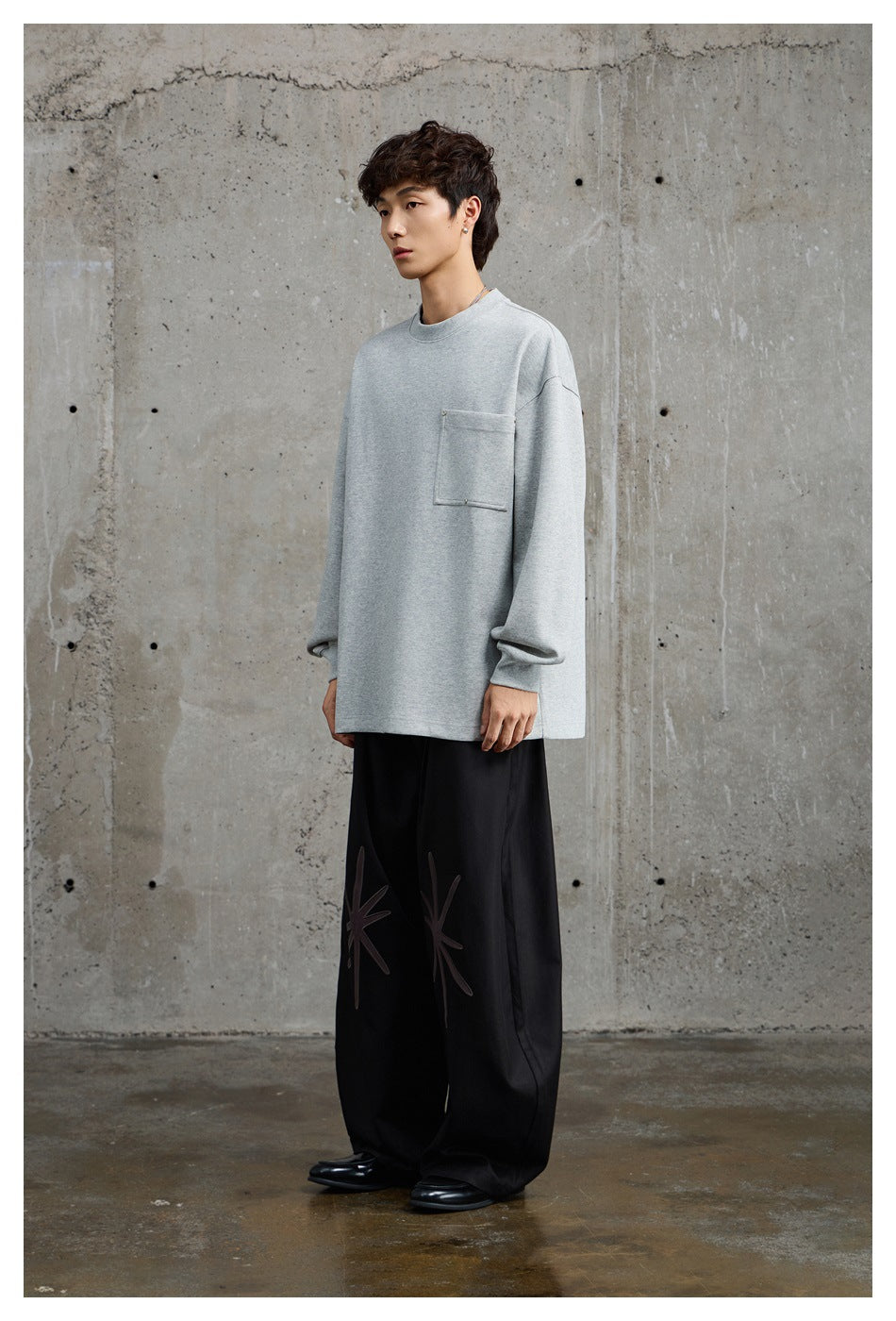 Model wearing an oversized light gray crewneck sweatshirt with a large chest pocket and loose black graphic trousers.