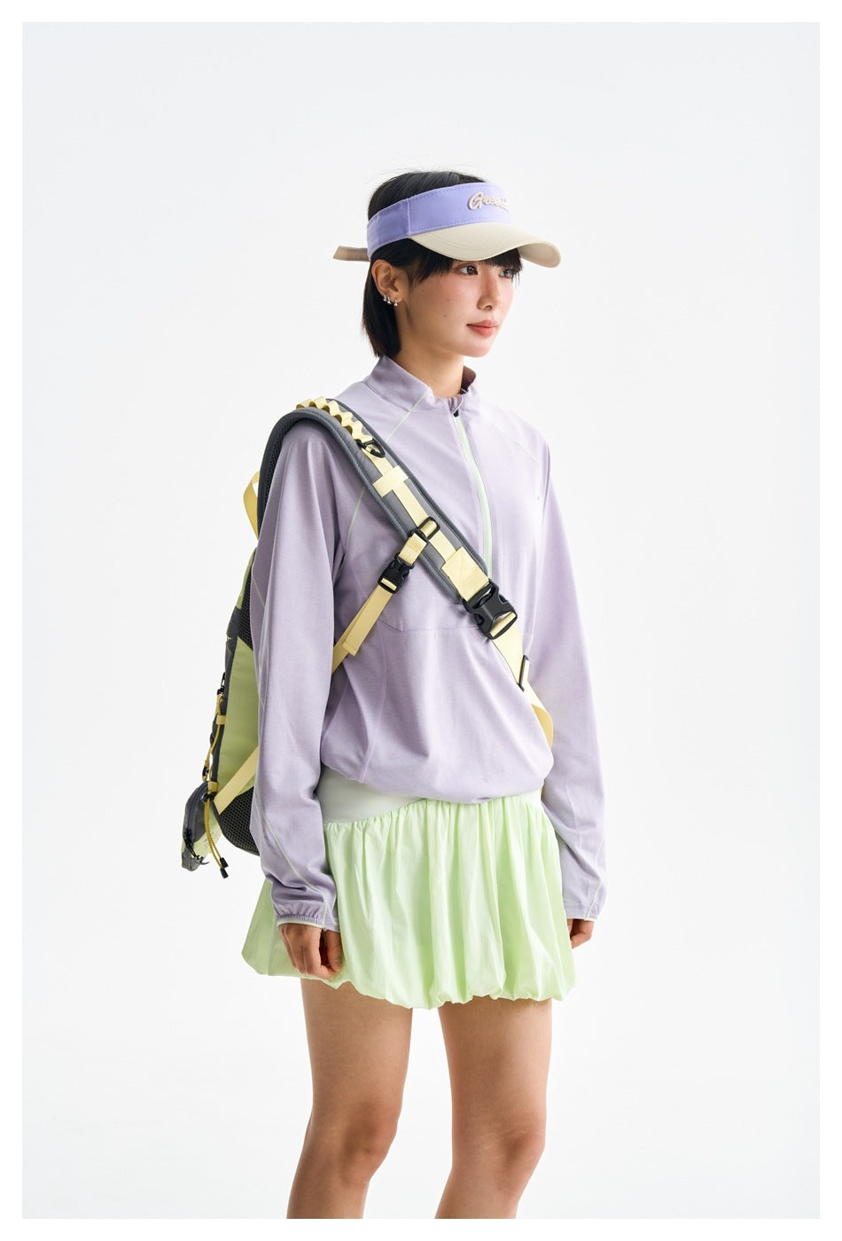 Model wearing a lilac quarter-zip top, pale mint bubble skirt and lavender visor, with a yellow and gray backpack.