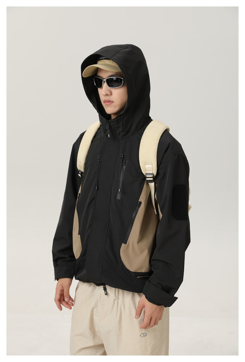 Model wearing the black and tan color-block jacket with its hood up, showing the front pockets and sleeve patch.