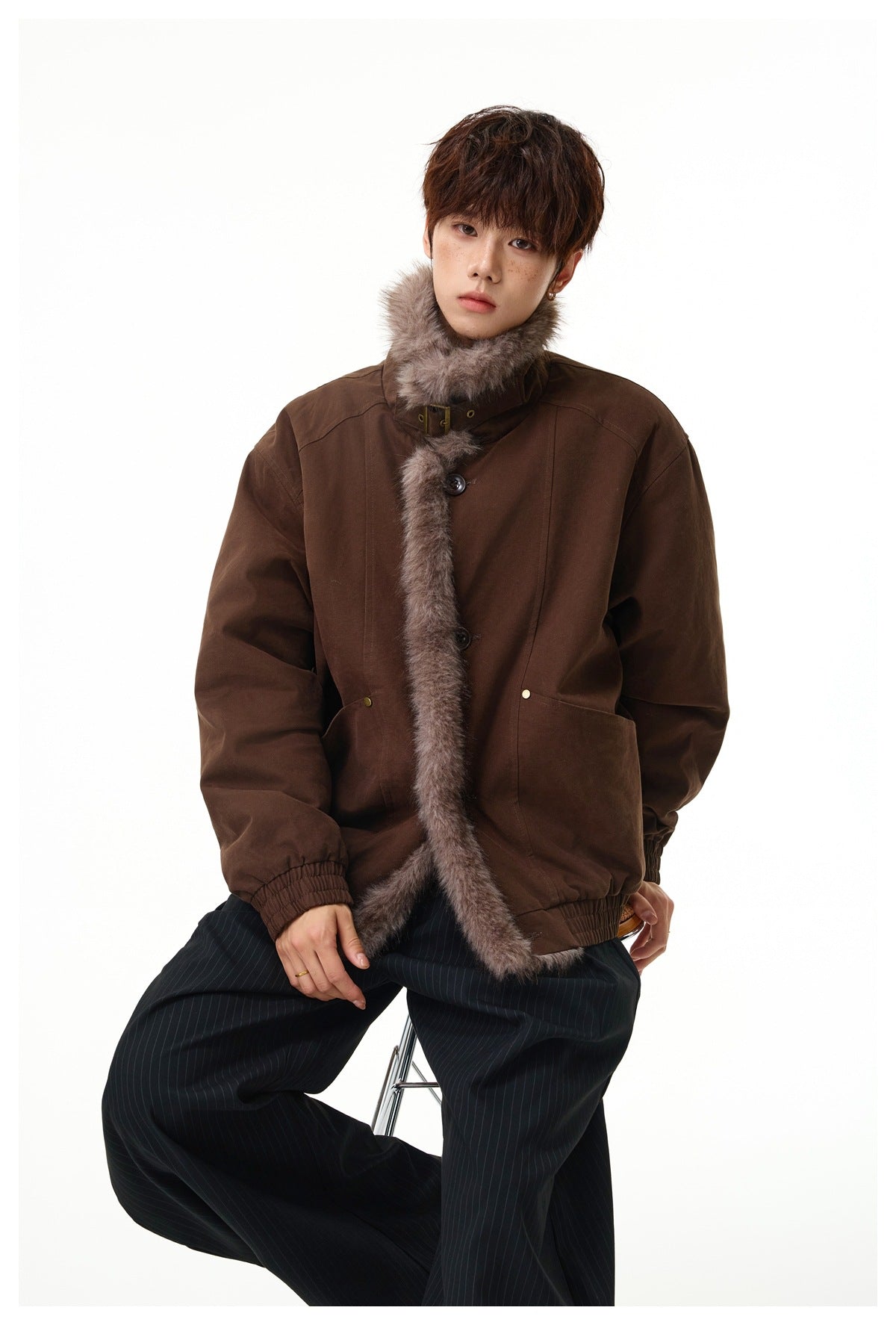 Model seated wearing the dark brown jacket with fluffy fleece-like collar and front trim over dark pinstripe wide pants