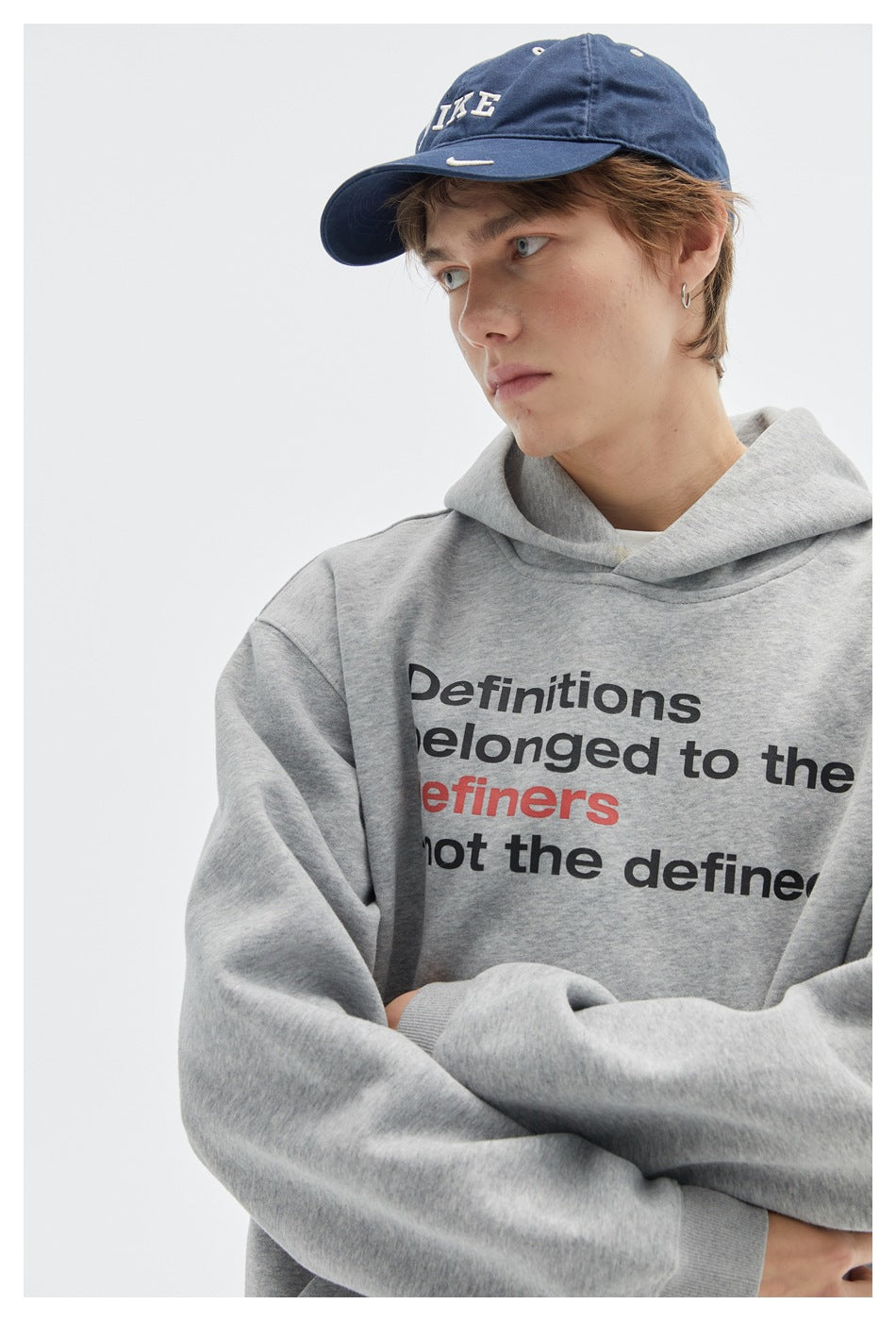 Close-up of a model in a light gray hoodie with black and red English text, wearing a navy cap.