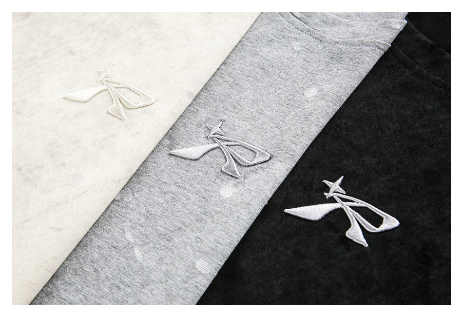 Close-up of a small embroidered motif on ivory, gray, and black distressed T-shirts.