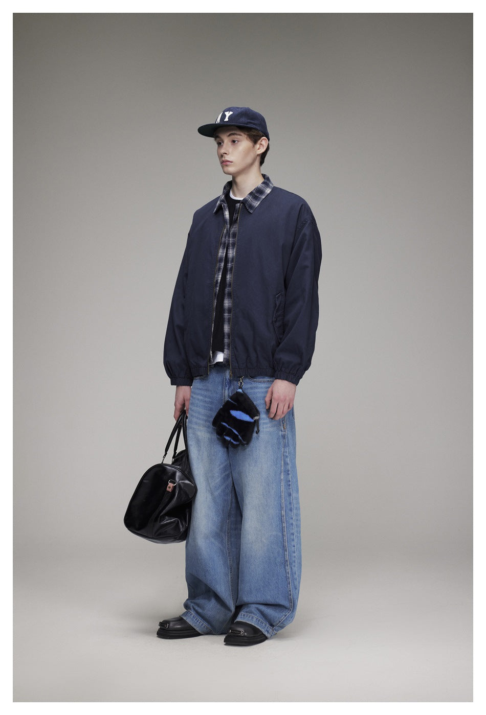 Model wearing navy zip jacket over a plaid shirt with wide-leg jeans, cap and black duffel bag