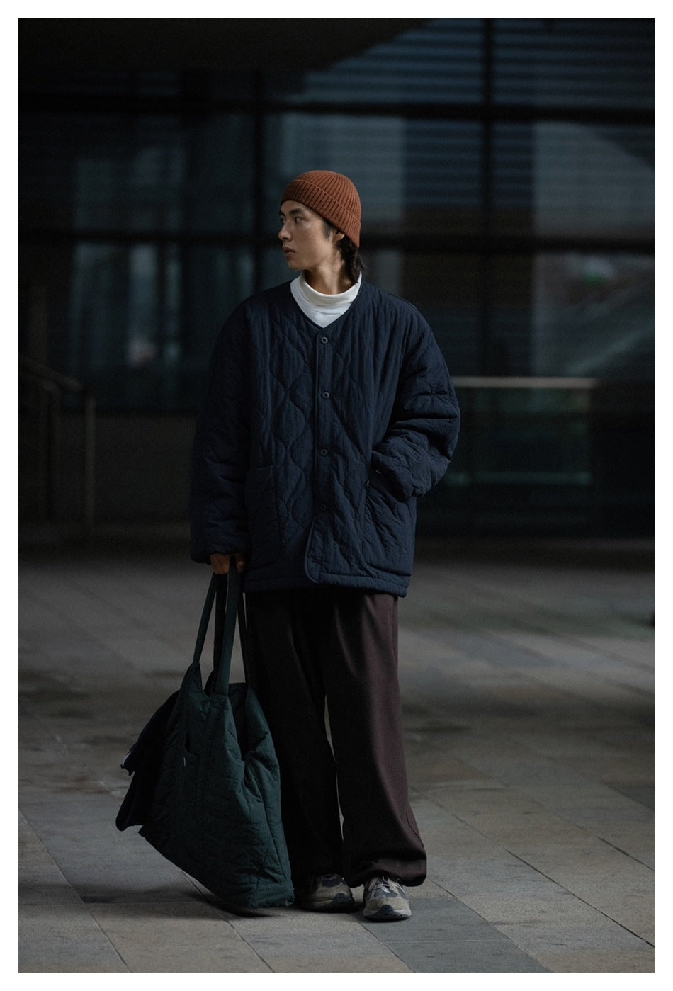 Model wearing a dark navy quilted V-neck jacket with buttons and patch pockets, styled with a rust beanie.