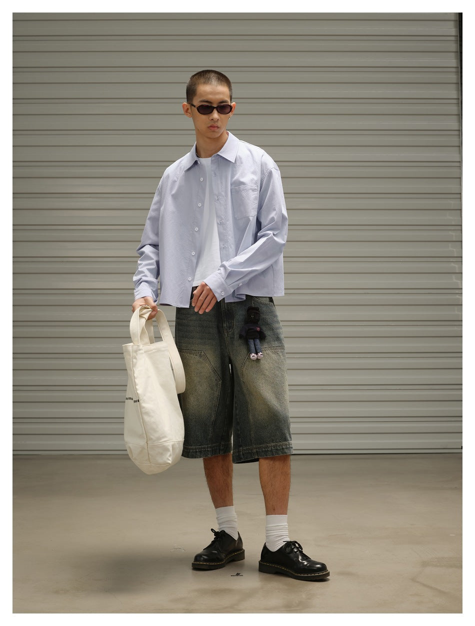 Three-quarter view of a model wearing a pale blue button-up shirt with a chest pocket and oversized dark denim shorts.