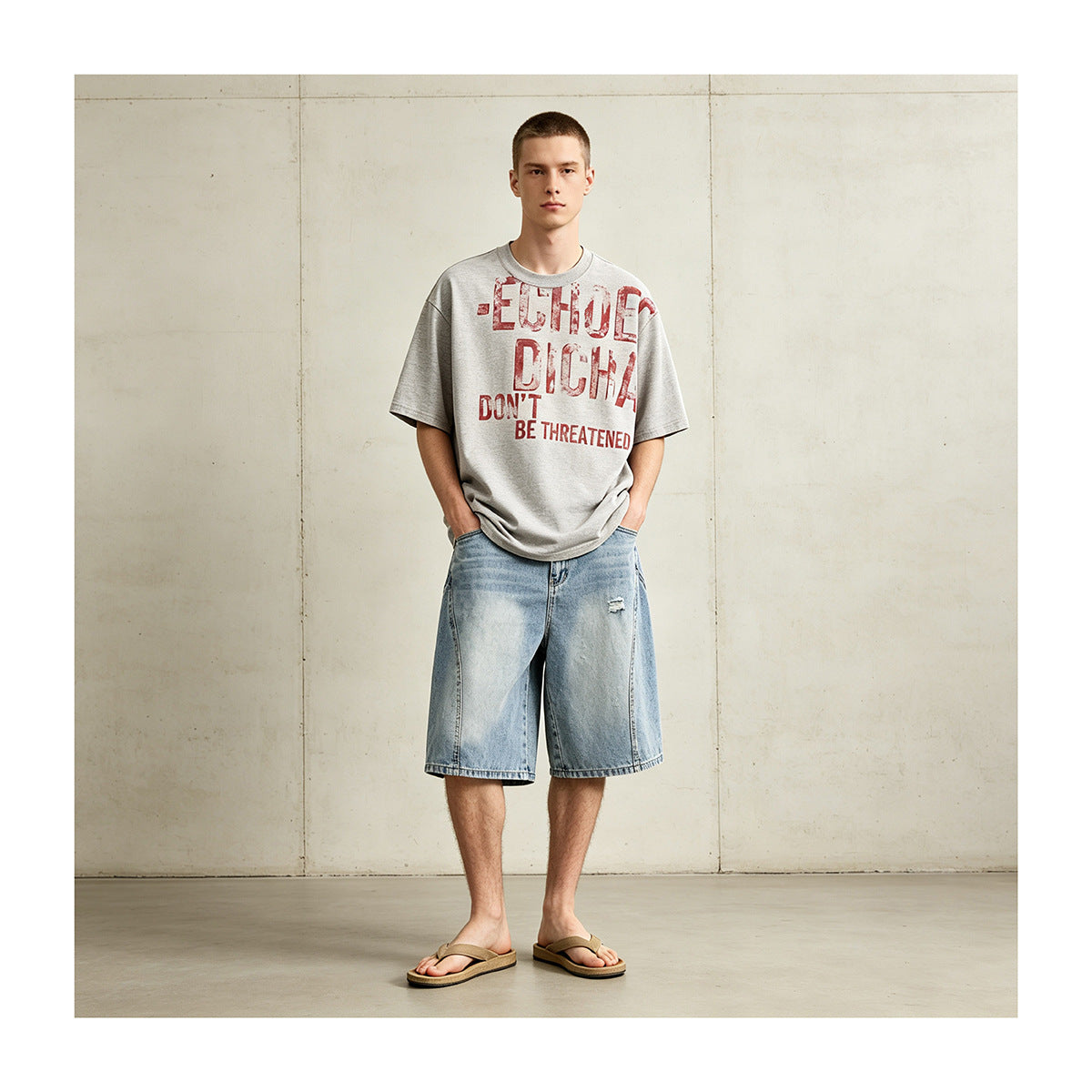 Man wearing light blue washed denim knee-length shorts with a gray graphic tee and sandals against a concrete wall