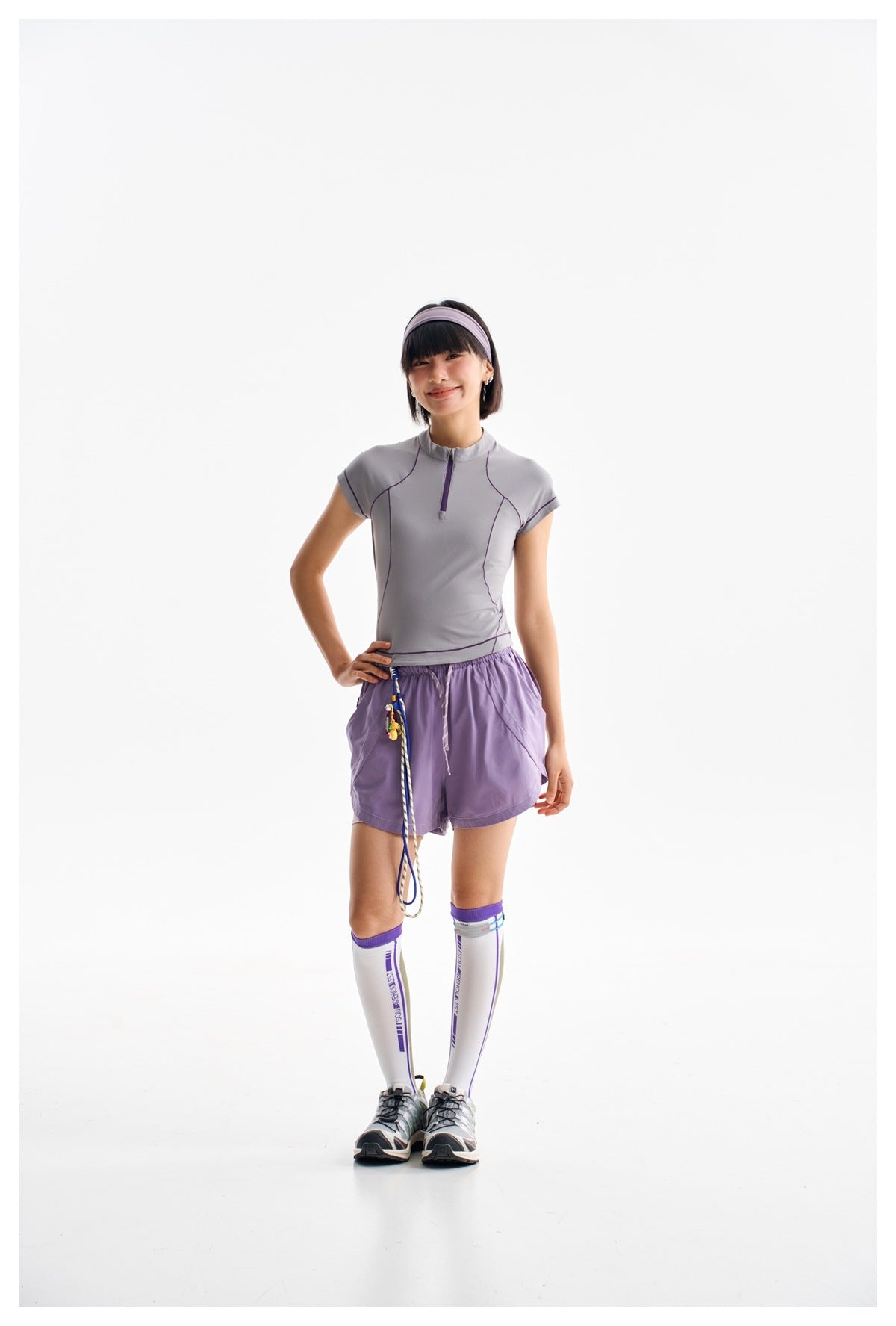 Model wearing a gray-lilac quarter-zip athletic top, purple shorts, and white knee-high socks with purple trim.