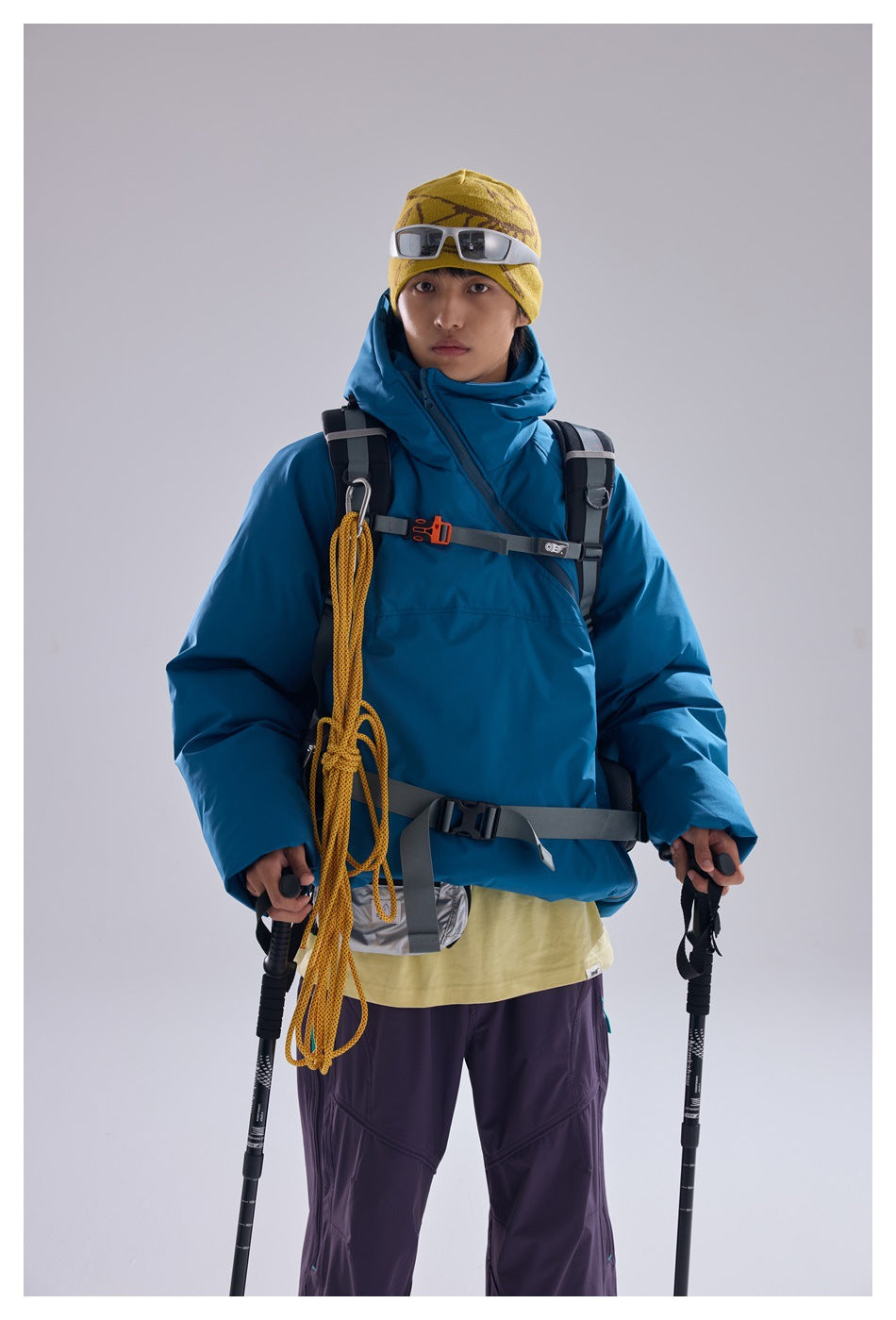 Front view of a model wearing a bright blue hooded jacket and purple pants with a yellow beanie and hiking gear.